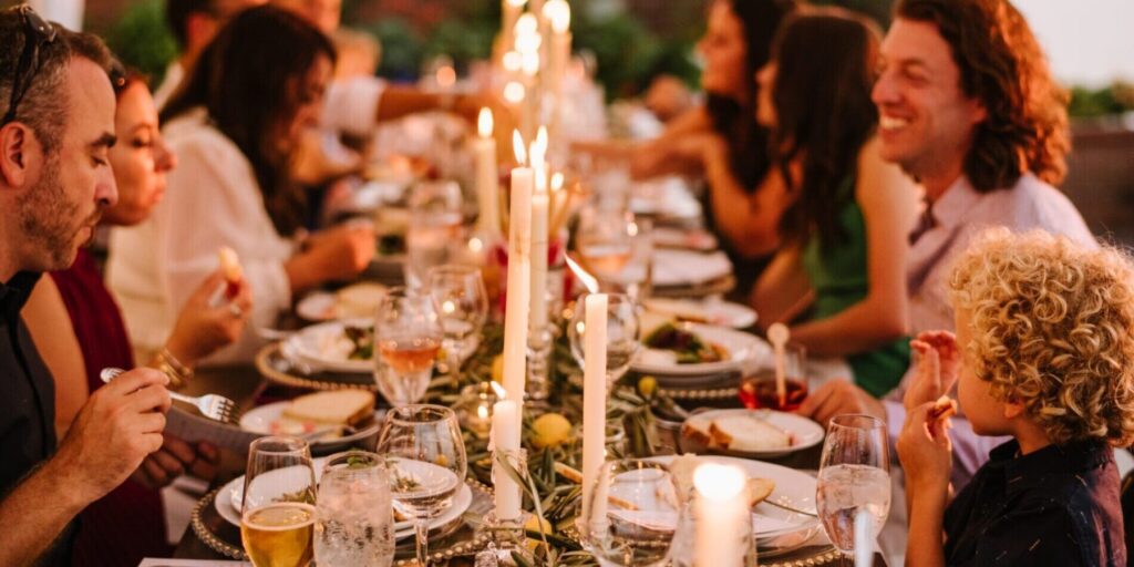 A group of people sit around a long, candlelit table sharing a meal, with plates of food, drinks, and glassware visible—capturing the warmth and elegance often found in wedding and event catering.