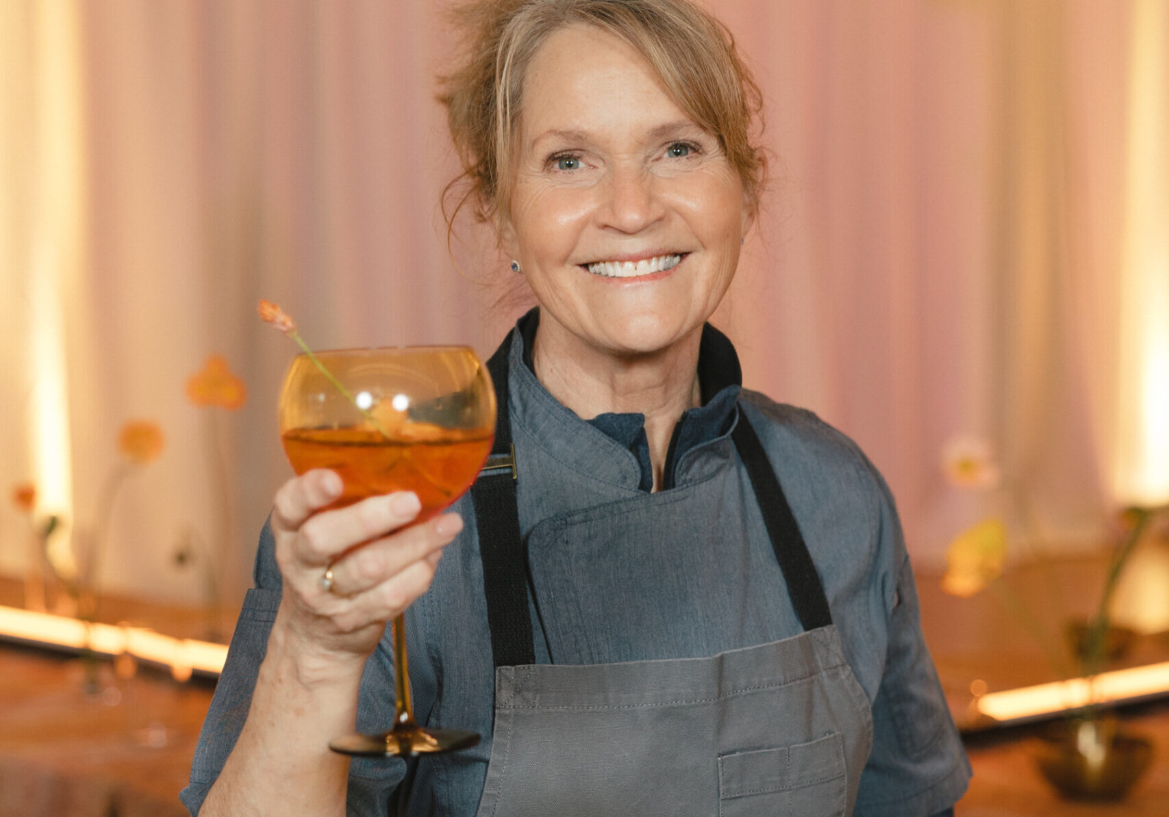 A woman in a gray apron and blue shirt smiles while holding up a cocktail in a glass, standing indoors with soft lighting—capturing the warm hospitality of About Lisa Dupar Catering Seattle.