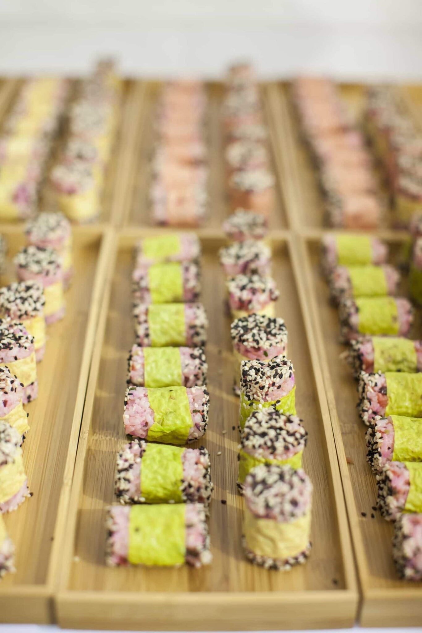 Rows of neatly arranged sushi rolls with green and pink rice, wrapped in seaweed and sprinkled with sesame seeds, displayed on wooden trays—perfect for catering for fundraisers.