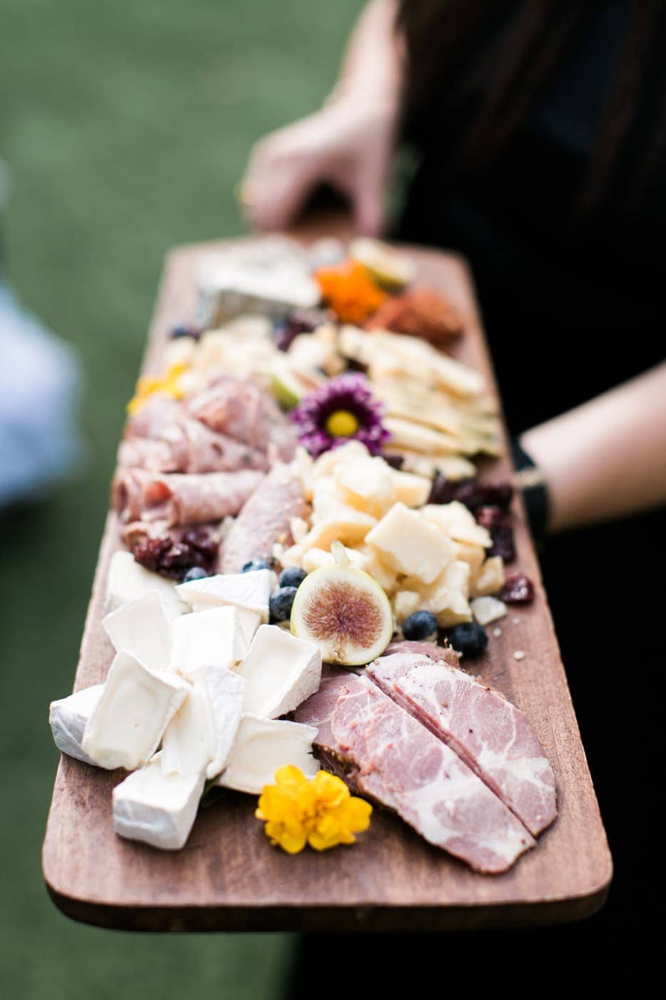 A person holds a wooden platter topped with assorted cheeses, cured meats, figs, blueberries, and edible flowers—perfect inspiration for elegant wedding catering Seattle style.
