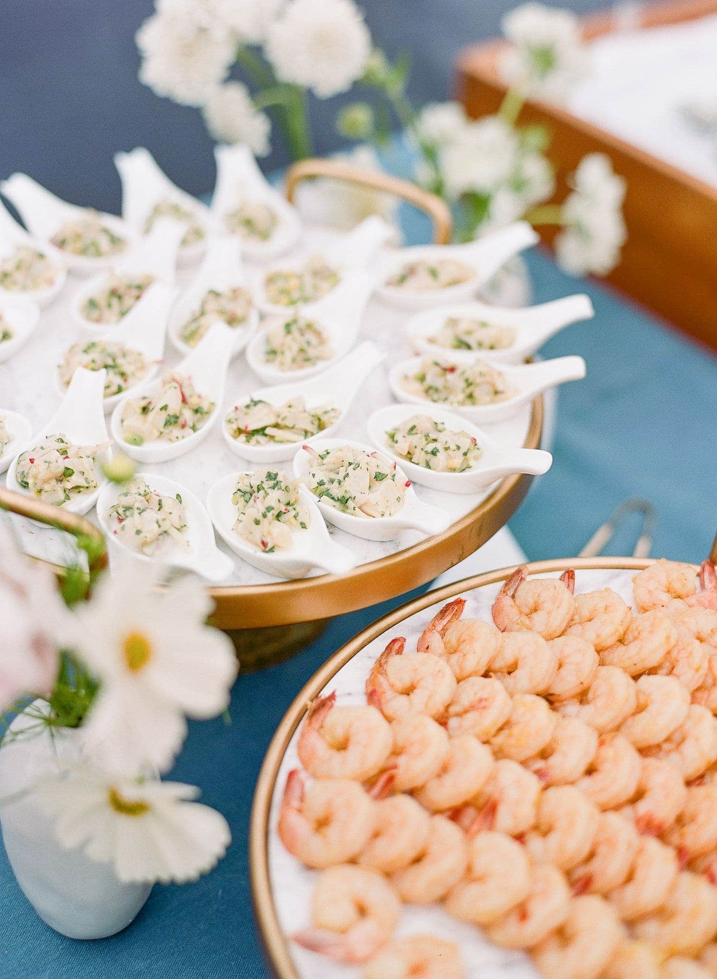 A platter of shrimp sits beside a tray of small white spoons filled with a chopped seafood appetizer, perfect for wedding catering Seattle, all arranged on a blue tablecloth with white flowers nearby.