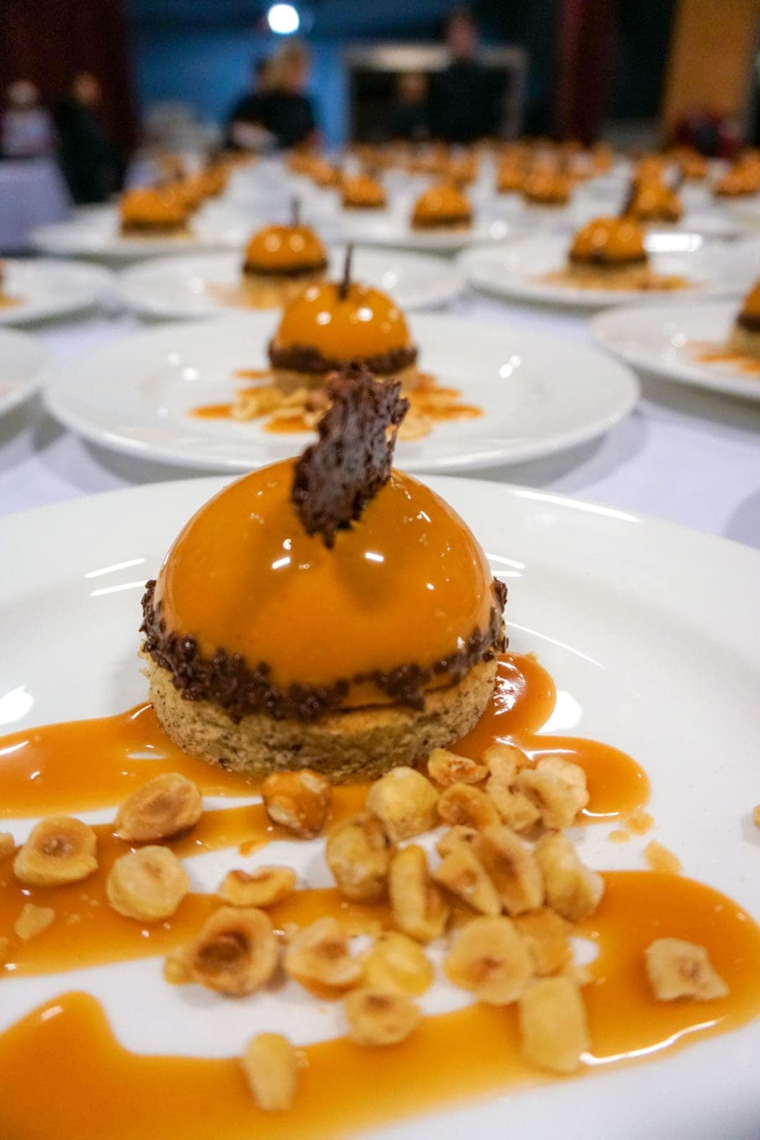 A plated dessert featuring a glossy orange mousse dome, garnished with chocolate, surrounded by caramel sauce and roasted hazelnuts—perfect for catering for fundraisers. Multiple plates are elegantly arranged in the background.