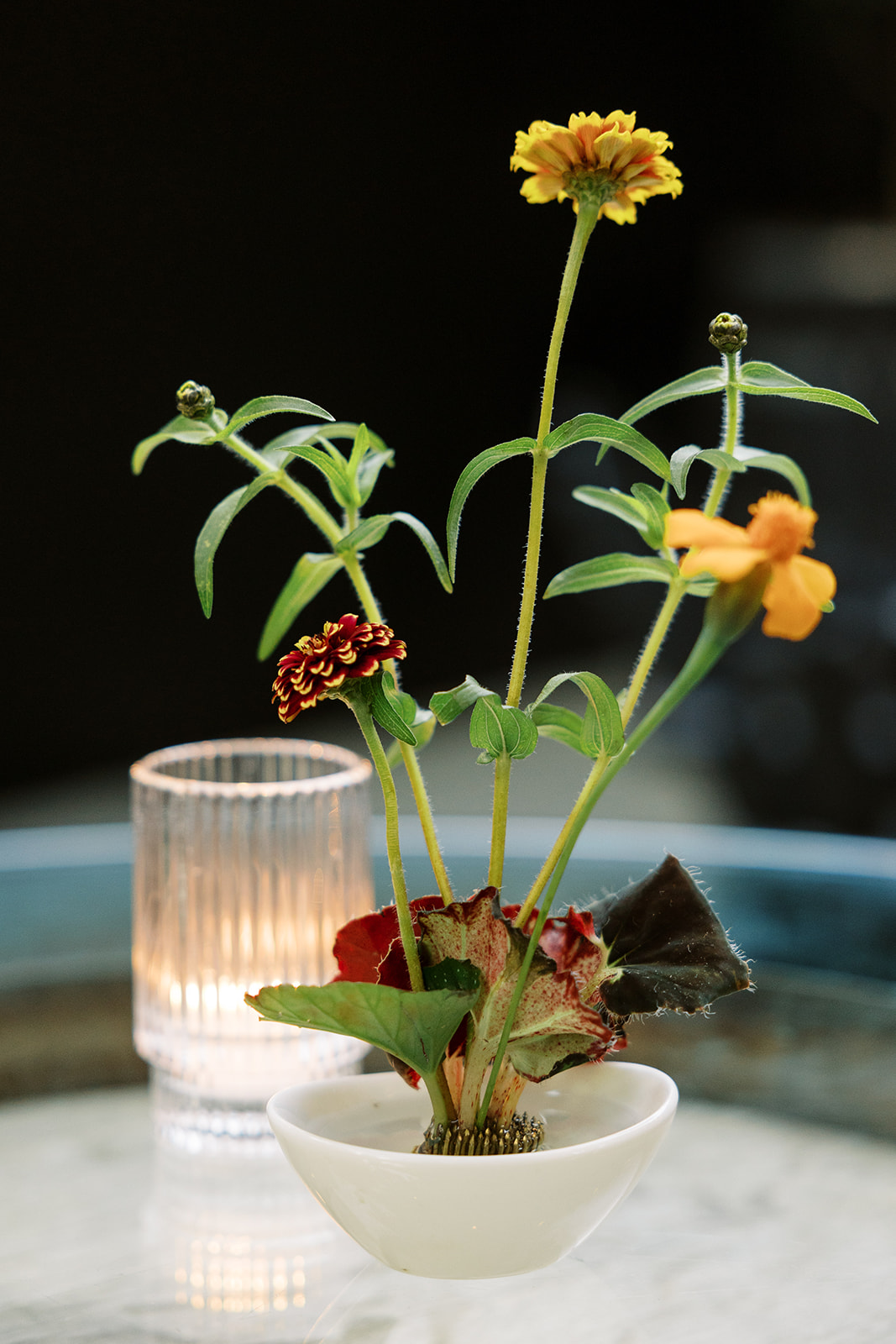 A small white bowl holds an arrangement of yellow and red flowers with green stems and leaves, set on a table beside a lit, ribbed glass candle—a tasteful touch perfect for catering for fundraisers.