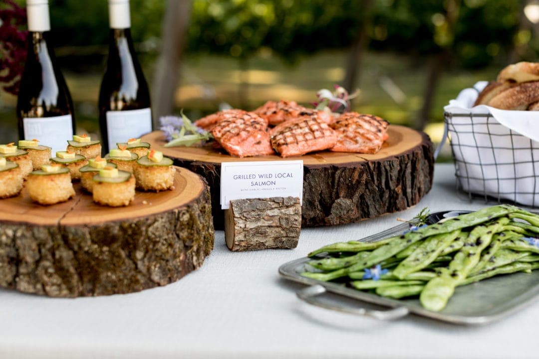 A table displays grilled wild salmon, fried appetizers with cucumber slices, a tray of grilled green beans, and bottles of wine outdoors—perfect for impressing guests at your next Corporate Catering Seattle event.