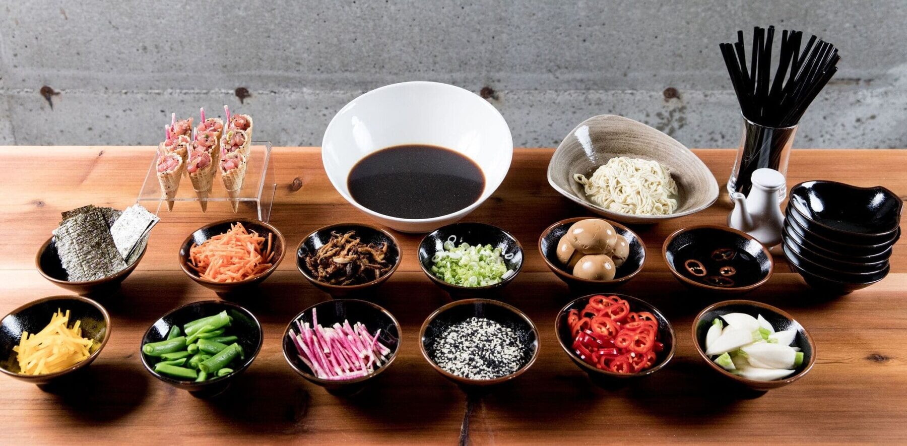 A variety of ramen ingredients in bowls, perfect for Corporate Catering Seattle, including vegetables, eggs, nori, noodles, and broth, are arranged neatly on a wooden table with black chopsticks and empty bowls nearby.