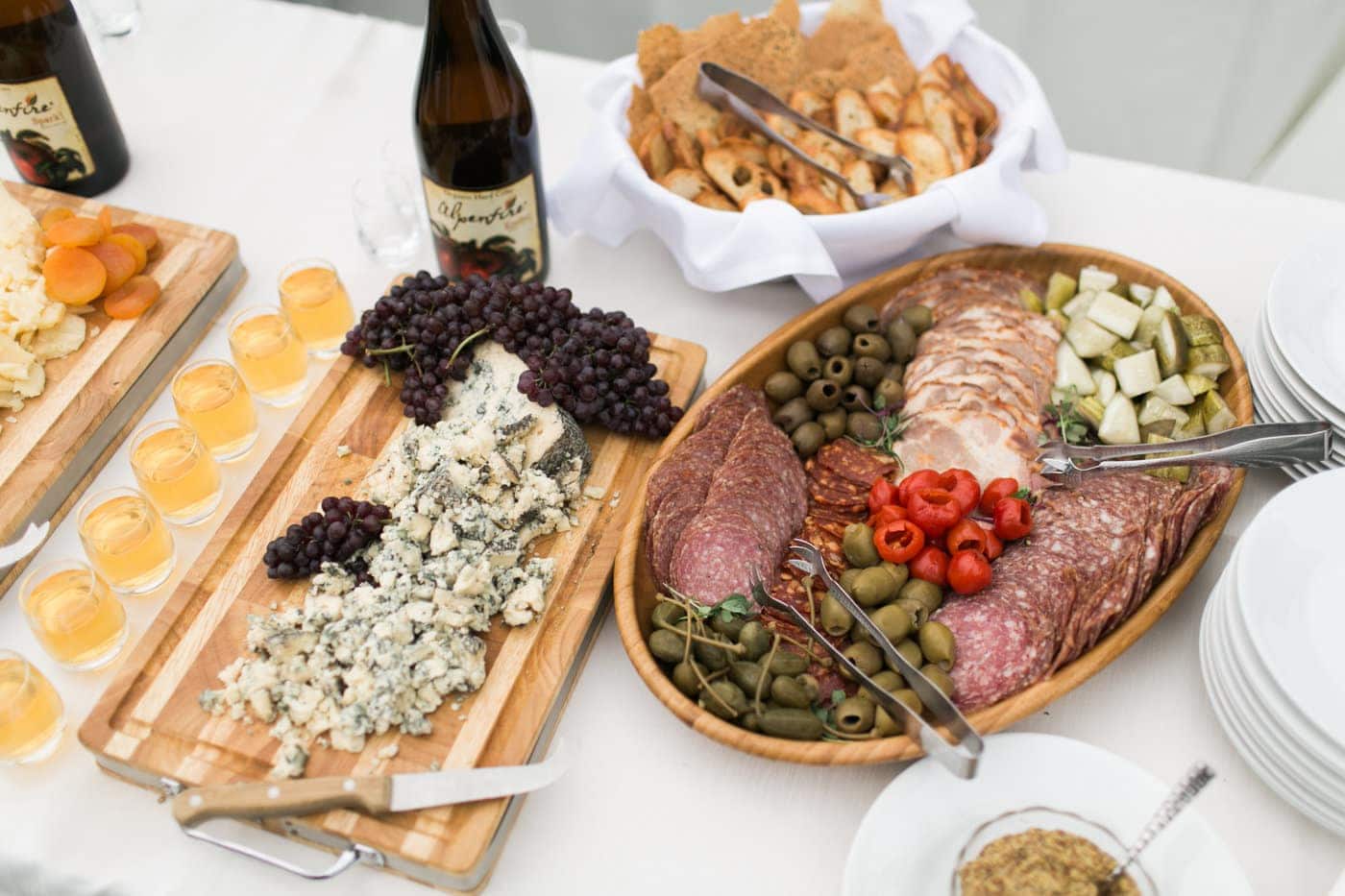 A table set for party catering with assorted cheeses, grapes, olives, sliced cured meats, crackers, small glasses of amber liquid, and bottles of wine.