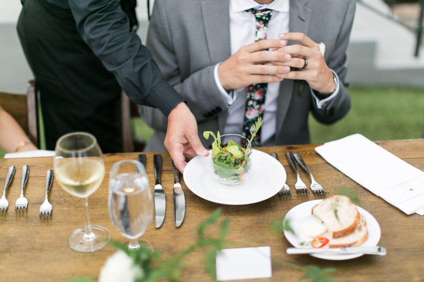 A person in a gray suit sits at a table as a server places a salad in front of them, with wine, water, bread, and utensils arranged neatly—showcasing the elegance and attention to detail of wedding catering Seattle has to offer.
