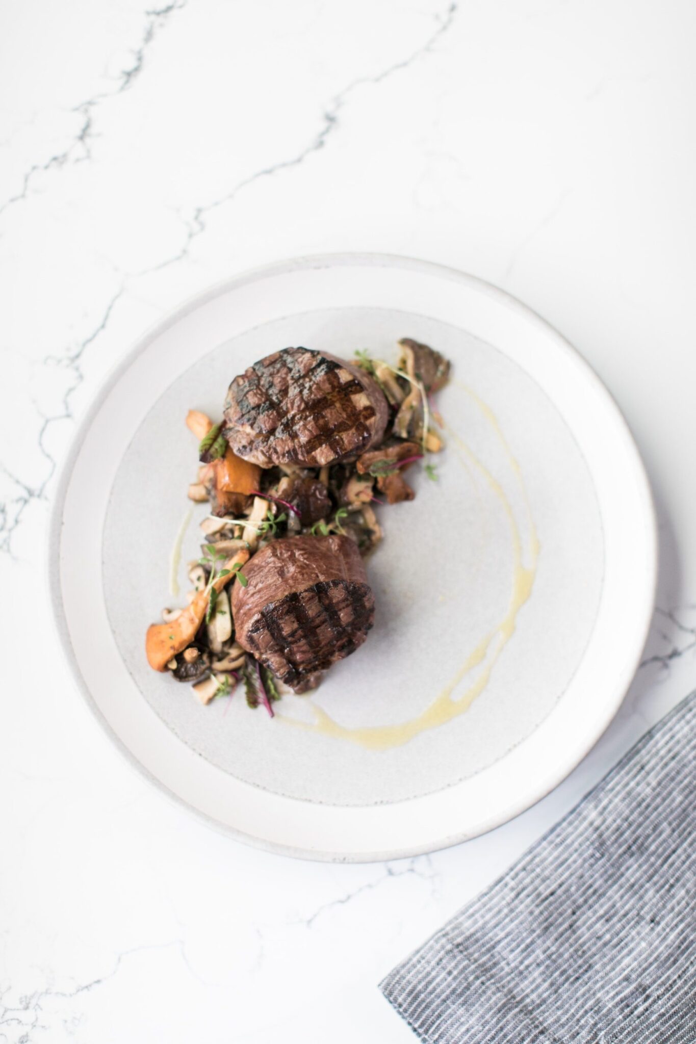 Two grilled steaks served on a white plate with sautéed vegetables and a light sauce, presented on a marble surface beside a striped napkin—perfect for an elevated Corporate Catering Seattle experience.