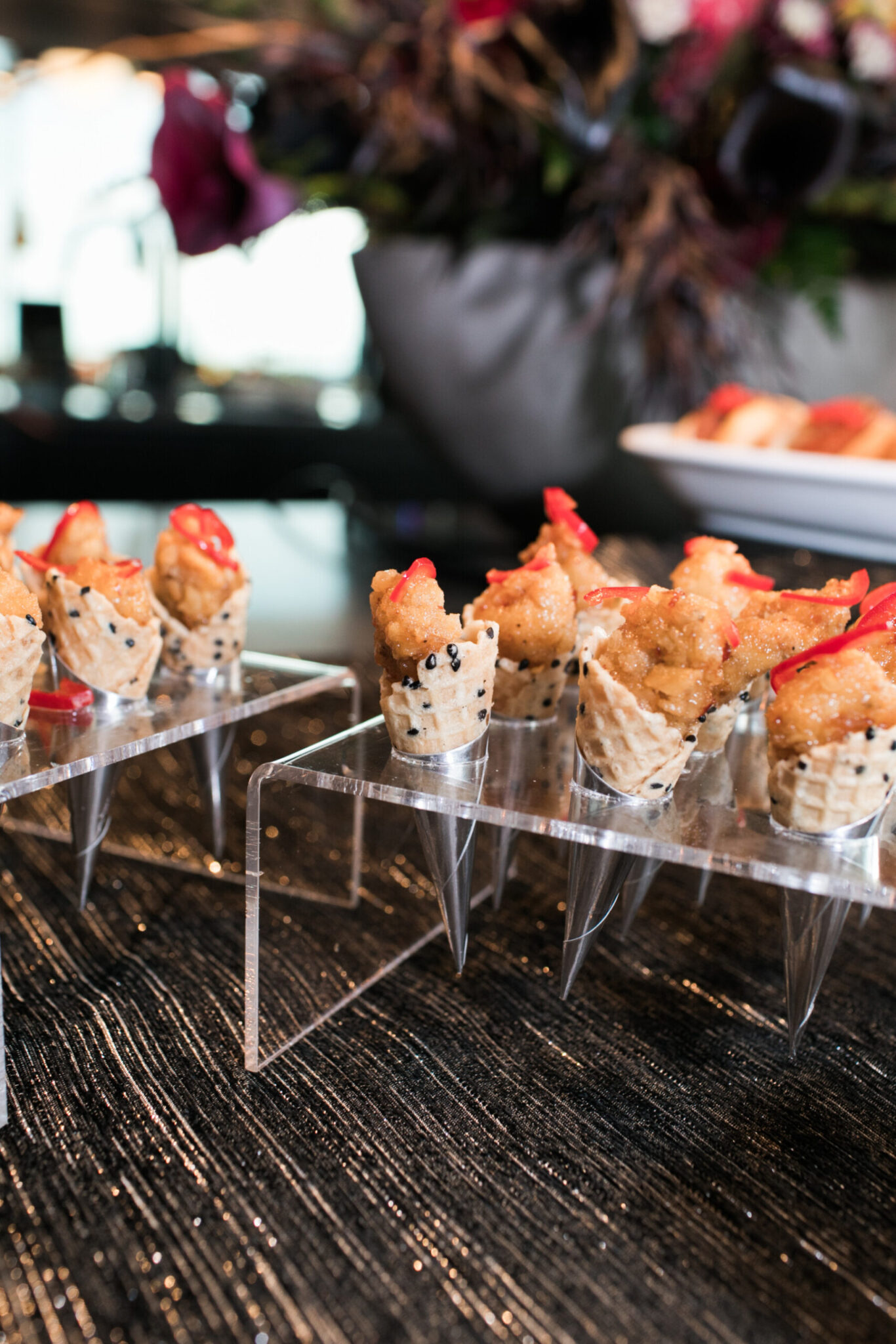 Miniature sushi cones topped with a slice of red pepper are displayed upright in clear acrylic holders on a dark textured tablecloth—perfect for elegant catering for fundraisers.