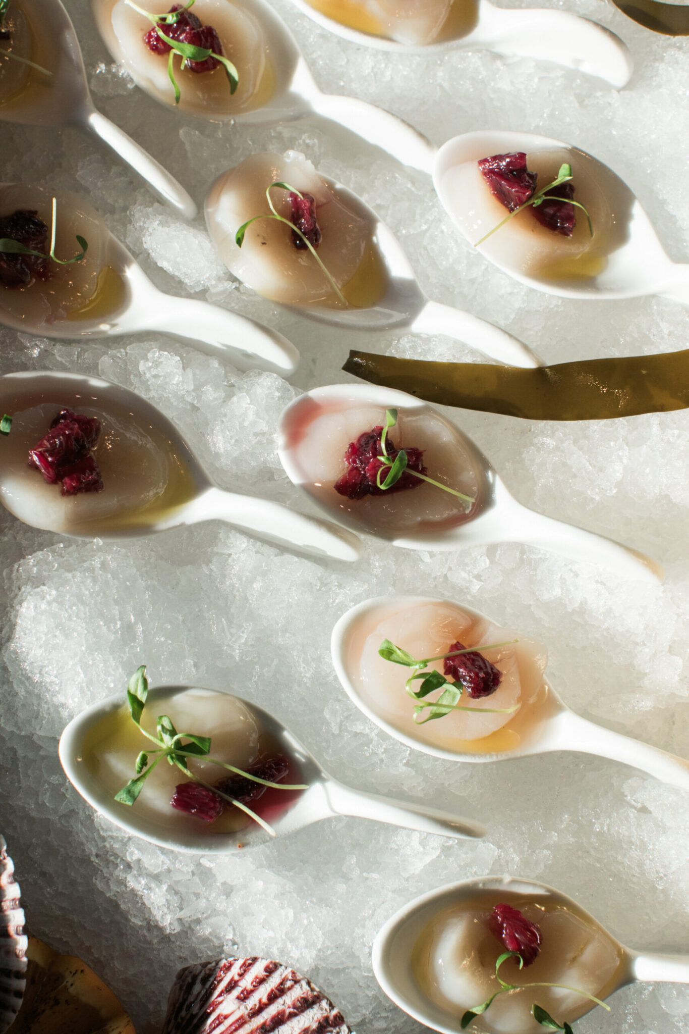 White ceramic spoons with raw scallops topped with red relish and microgreens are arranged on crushed ice—perfect for elegant wedding catering in Seattle.