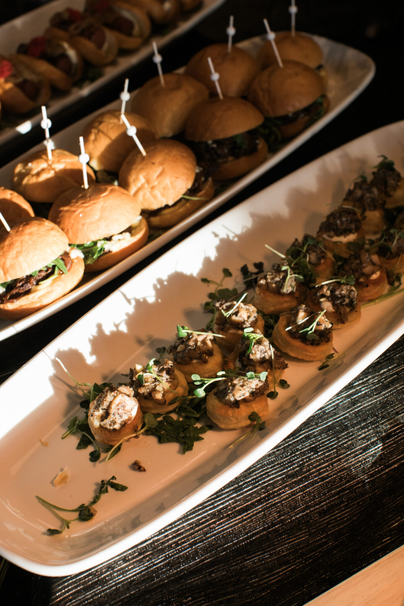 Two white trays of appetizers for party catering rest on a dark tablecloth; one features mini sandwiches, while the other offers small bread rounds topped with a mushroom mixture and fresh greens.