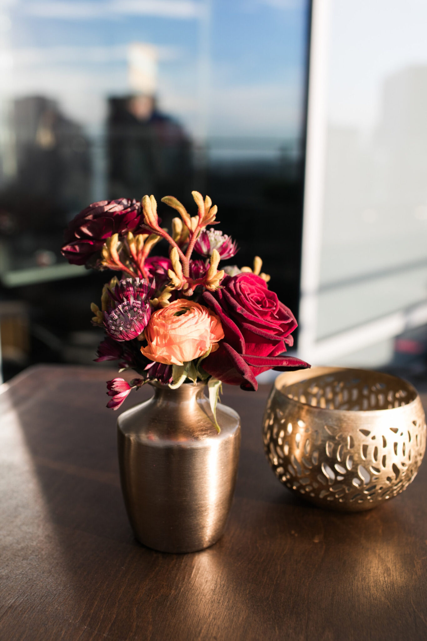 A small bronze vase with red and orange flowers sits on a wooden table next to a decorative gold candle holder, creating an inviting setting perfect for party catering.