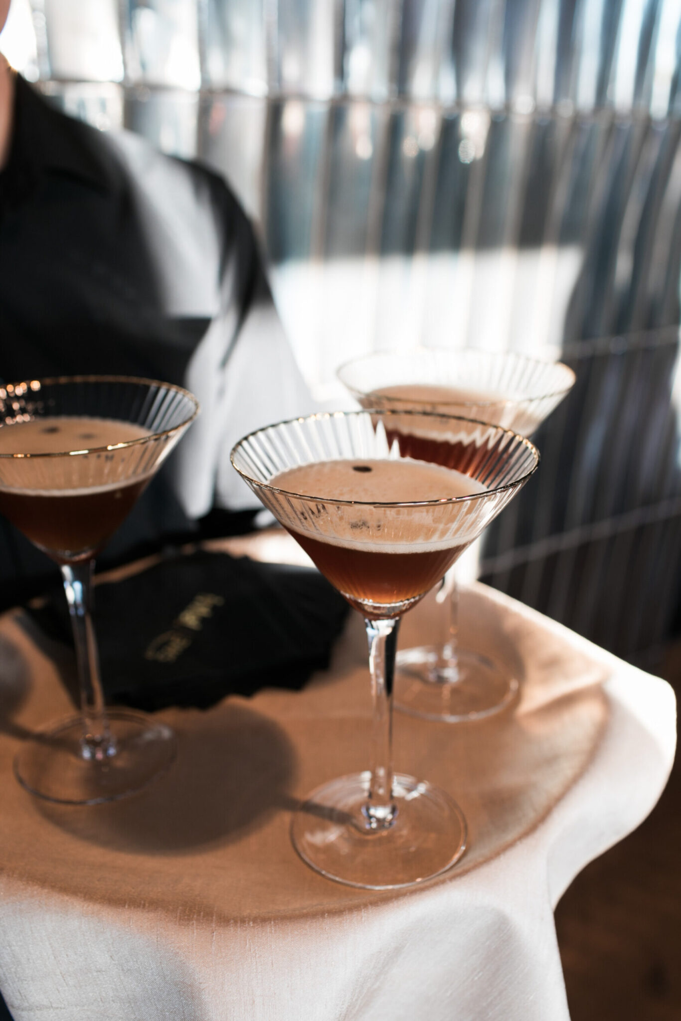 Three espresso martinis in coupe glasses sit on a white tablecloth, with a black napkin nearby—perfect for adding a touch of elegance to your next party catering event.