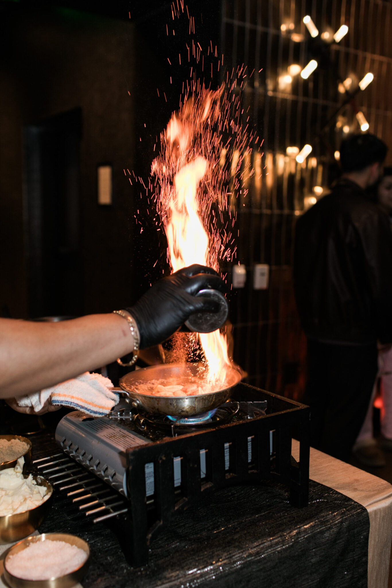 A person wearing a black glove pours liquid into a pan on a portable stove, causing flames to rise, while preparing ingredients for catering for fundraisers.