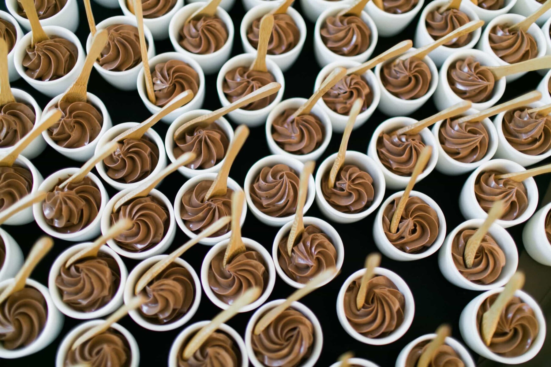 Rows of small white cups filled with swirled chocolate mousse, each with a wooden spoon inserted, arranged closely together on a black surface—perfect for catering for fundraisers or elegant events.