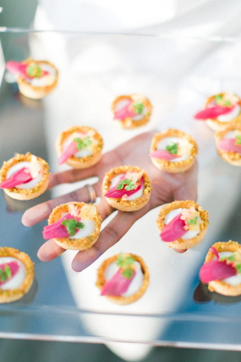 A hand holds a clear tray with several small, round appetizers topped with cream, herbs, and pink garnish—perfect bites for Corporate Catering Seattle events.