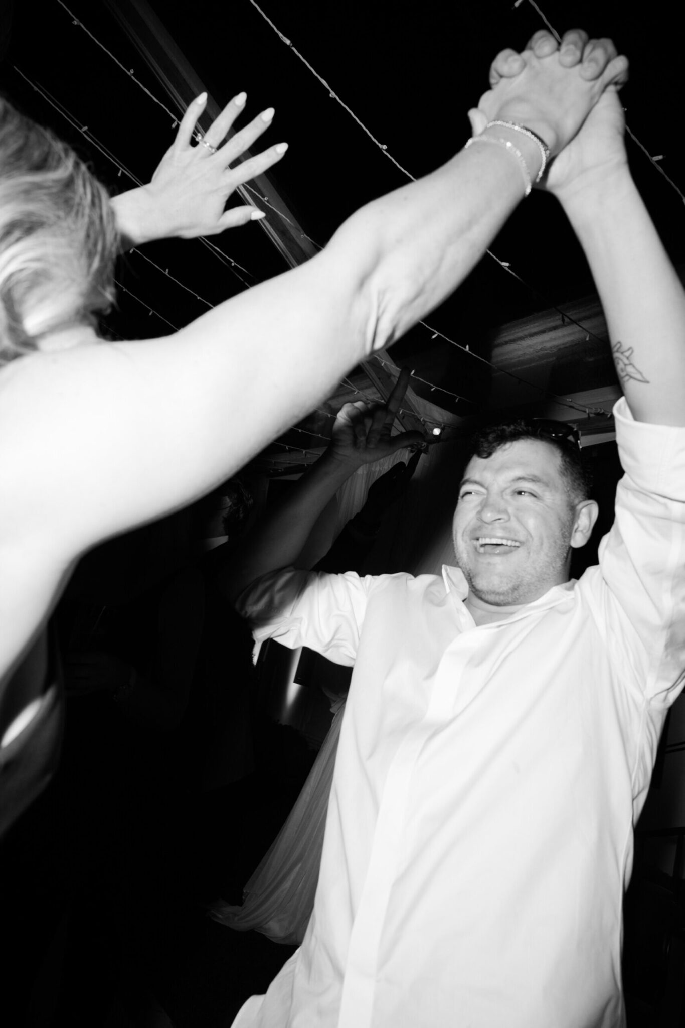 A man in a white shirt smiles and raises his arms while dancing with another person under string lights at a wedding catering Seattle event in a dark setting.