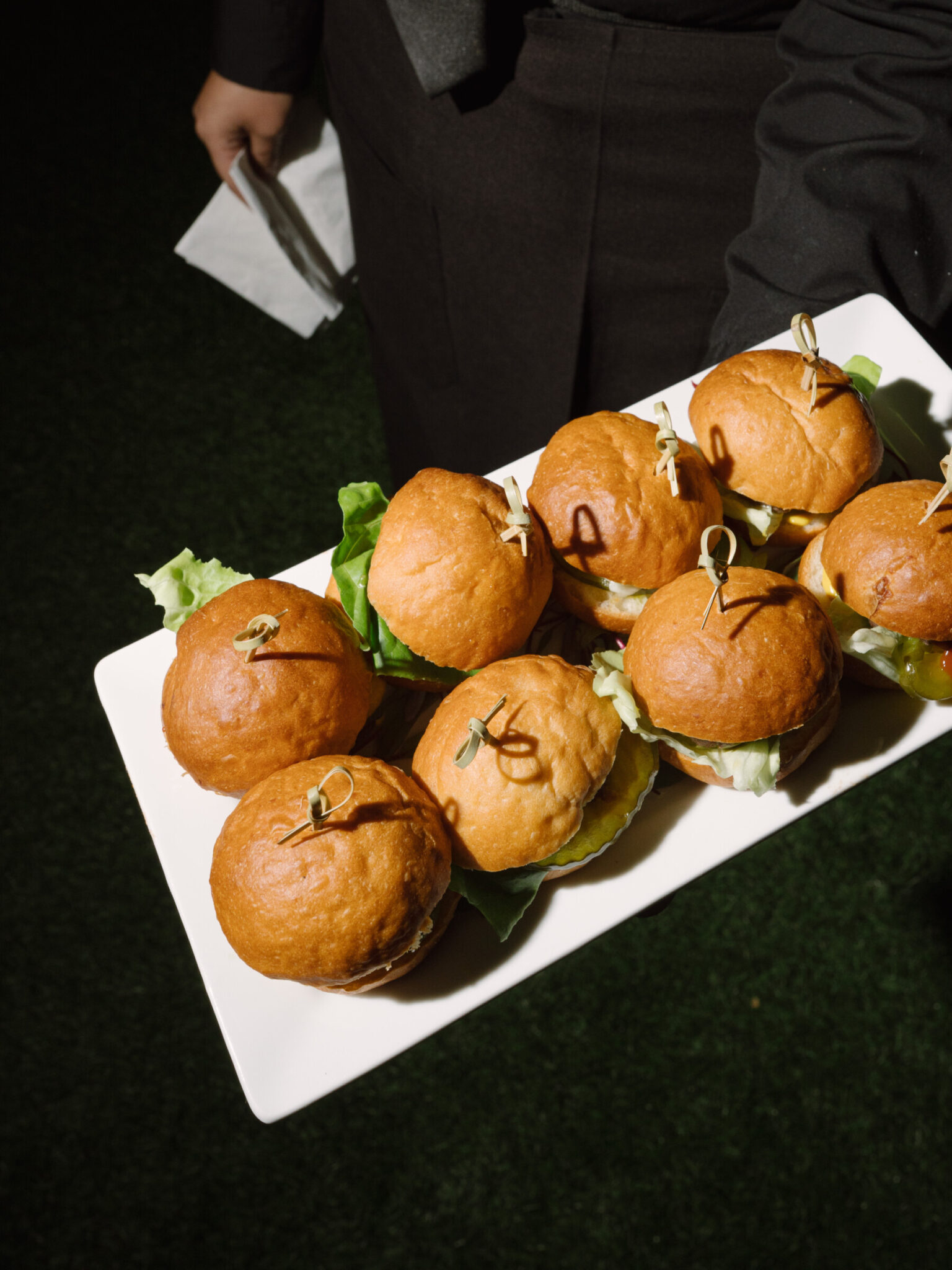 A person holding a white rectangular plate with seven mini burgers secured with toothpicks, served on buns with lettuce—perfect for corporate catering in Seattle.