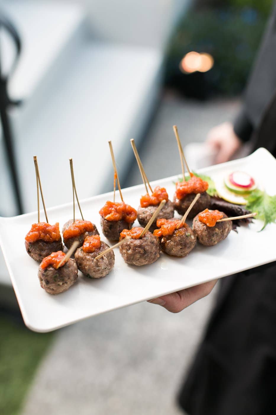 A hand holds a rectangular white plate with meatballs on skewers, each topped with red sauce and garnished with greens and sliced vegetables—perfect for party catering.
