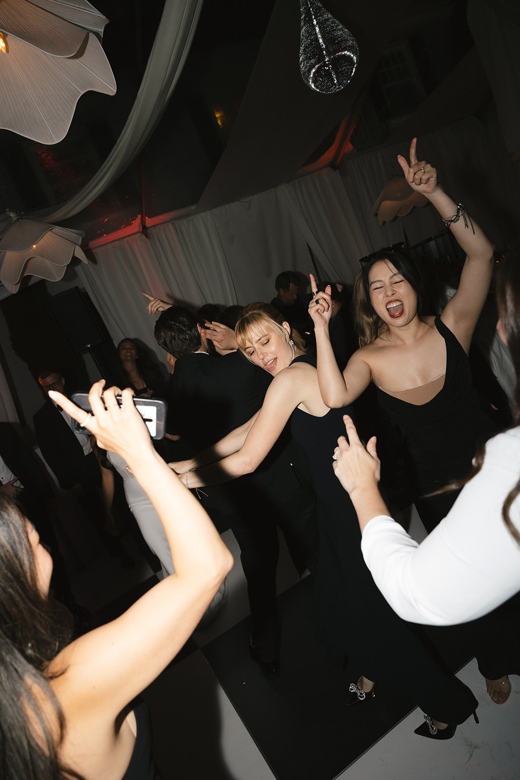 People dancing and celebrating at an indoor event, with several individuals in black dresses and arms raised, surrounded by draped fabric and dim lighting—an unforgettable night elevated by top-notch wedding catering Seattle.