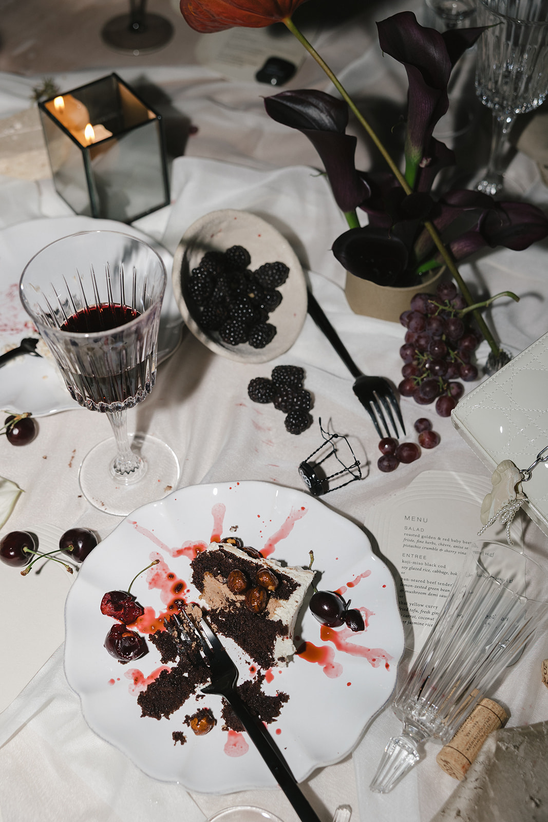 A partially eaten slice of chocolate cake on a white plate with cherries, berries, a wine glass, spilled drink, candle, and flowers on a white tablecloth—an elegant scene from wedding catering Seattle couples will love.