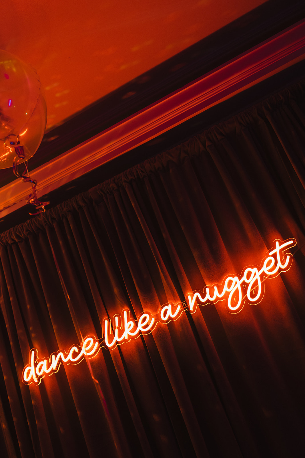 Neon orange sign with cursive letters reads "dance like a nugget," hanging against dark pleated curtains—perfect quirky decor inspiration for your wedding catering Seattle event. A balloon with a string is partly visible in the top left corner.