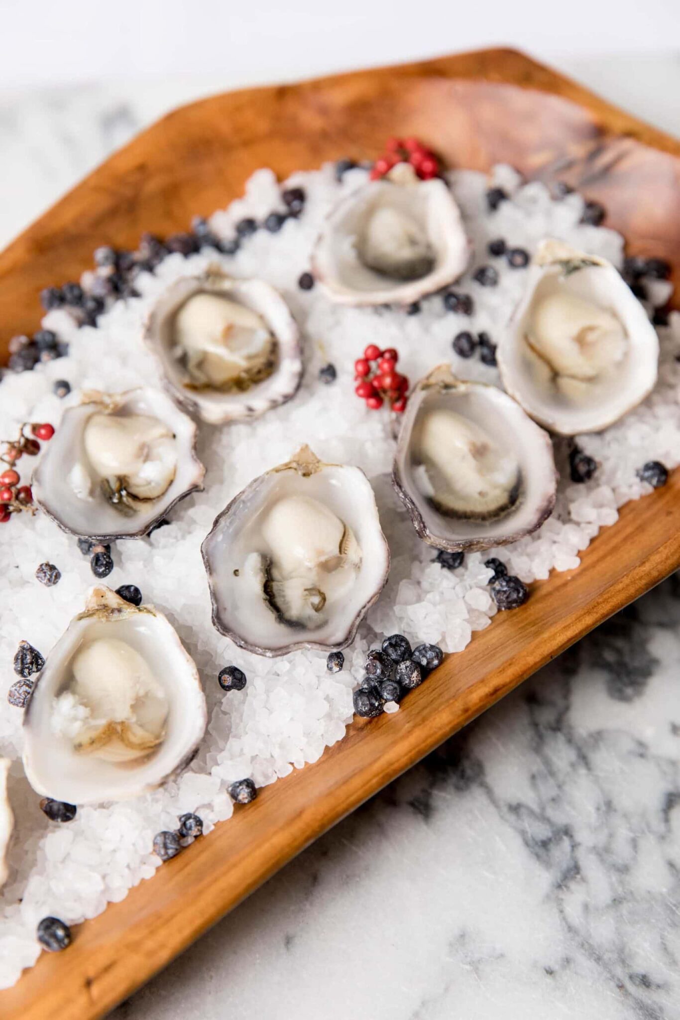 A wooden tray with half-shell oysters on a bed of coarse salt, garnished with black and red berries, perfect for elegant party catering, placed on a marble surface.