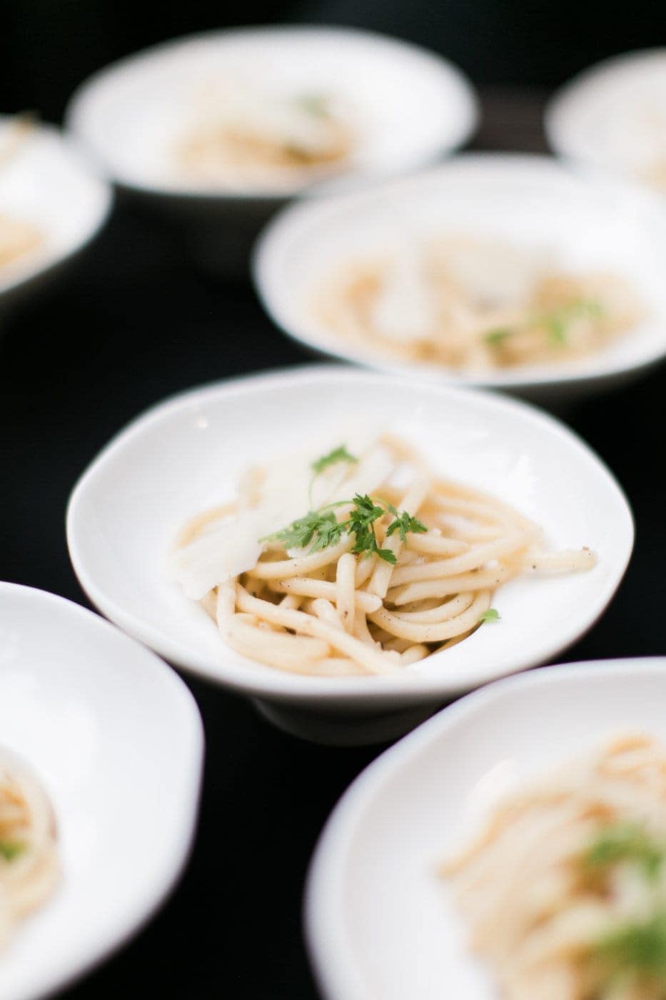 Small white bowls filled with pasta, garnished with herbs and cheese slices, are arranged on a dark surface—perfect for elegant Corporate Catering Seattle events.