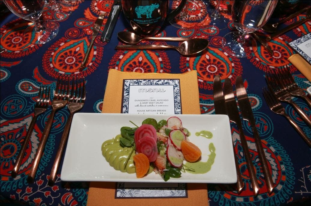 A rectangular plate with sliced avocado, radish, beet, and carrot is served on an orange napkin with a menu—perfect for catering for fundraisers—surrounded by silverware and glasses on a colorful patterned tablecloth.