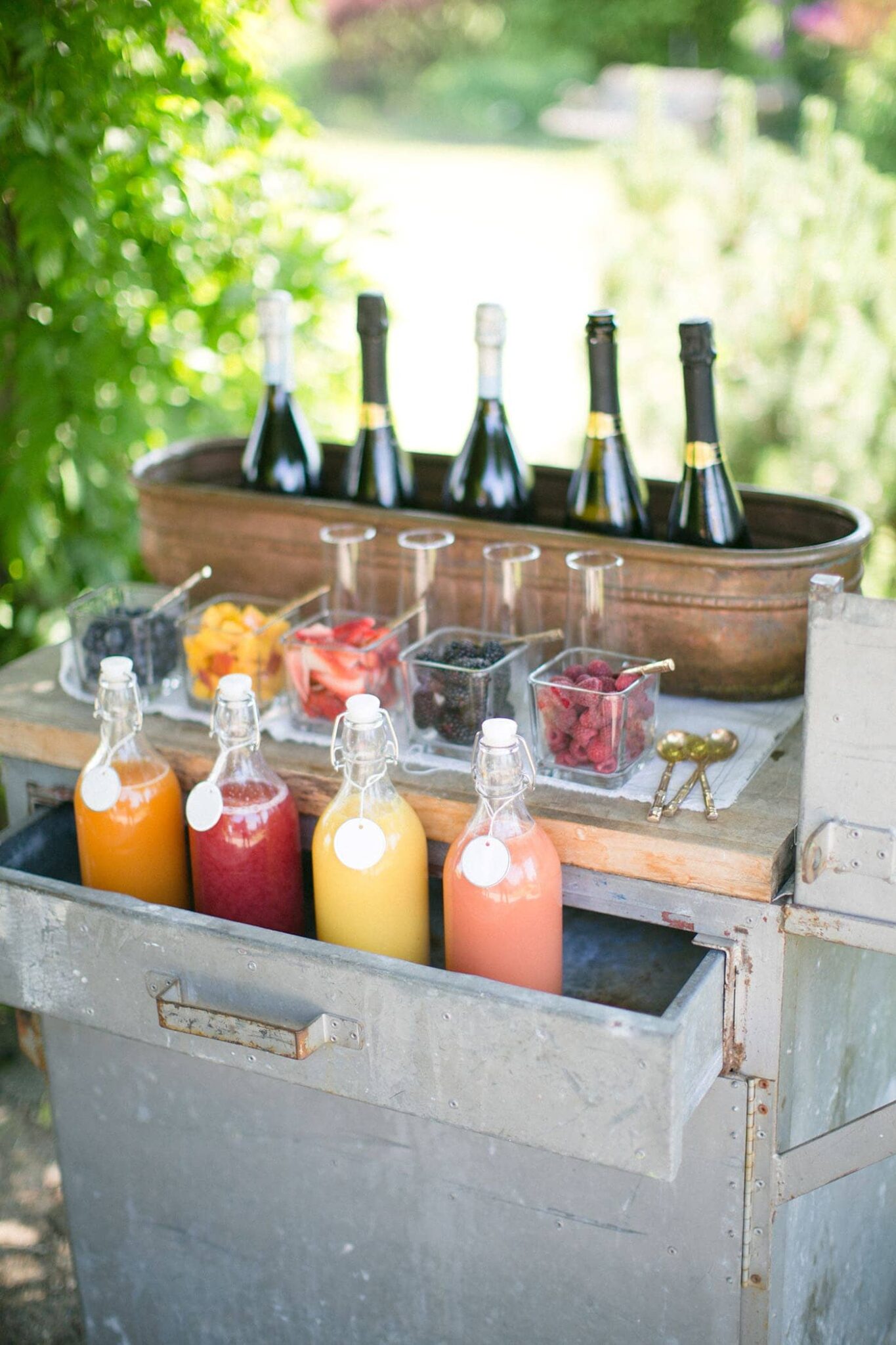 A party catering beverage station with bottles of juice, containers of fruit, several wine or champagne bottles, and serving utensils is set on a metal cart outdoors.