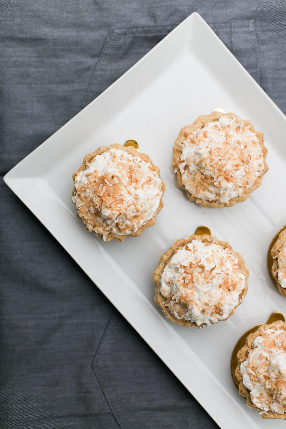 Five mini tarts topped with toasted coconut and cream are arranged on a rectangular white plate with a gray fabric background, making them perfect for stylish party catering.