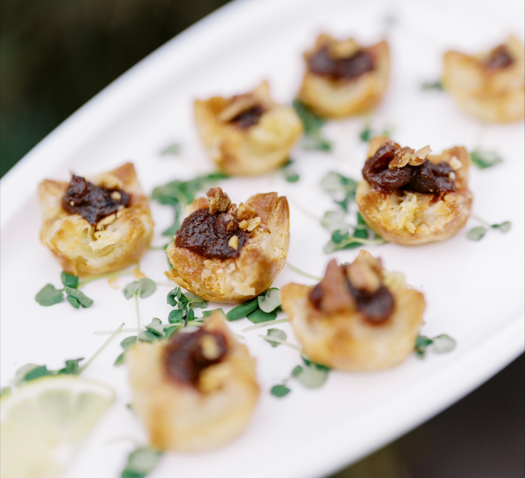 A white platter with several bite-sized puff pastry appetizers, perfect for catering for fundraisers, topped with a dark spread and small garnish, arranged on a bed of microgreens.