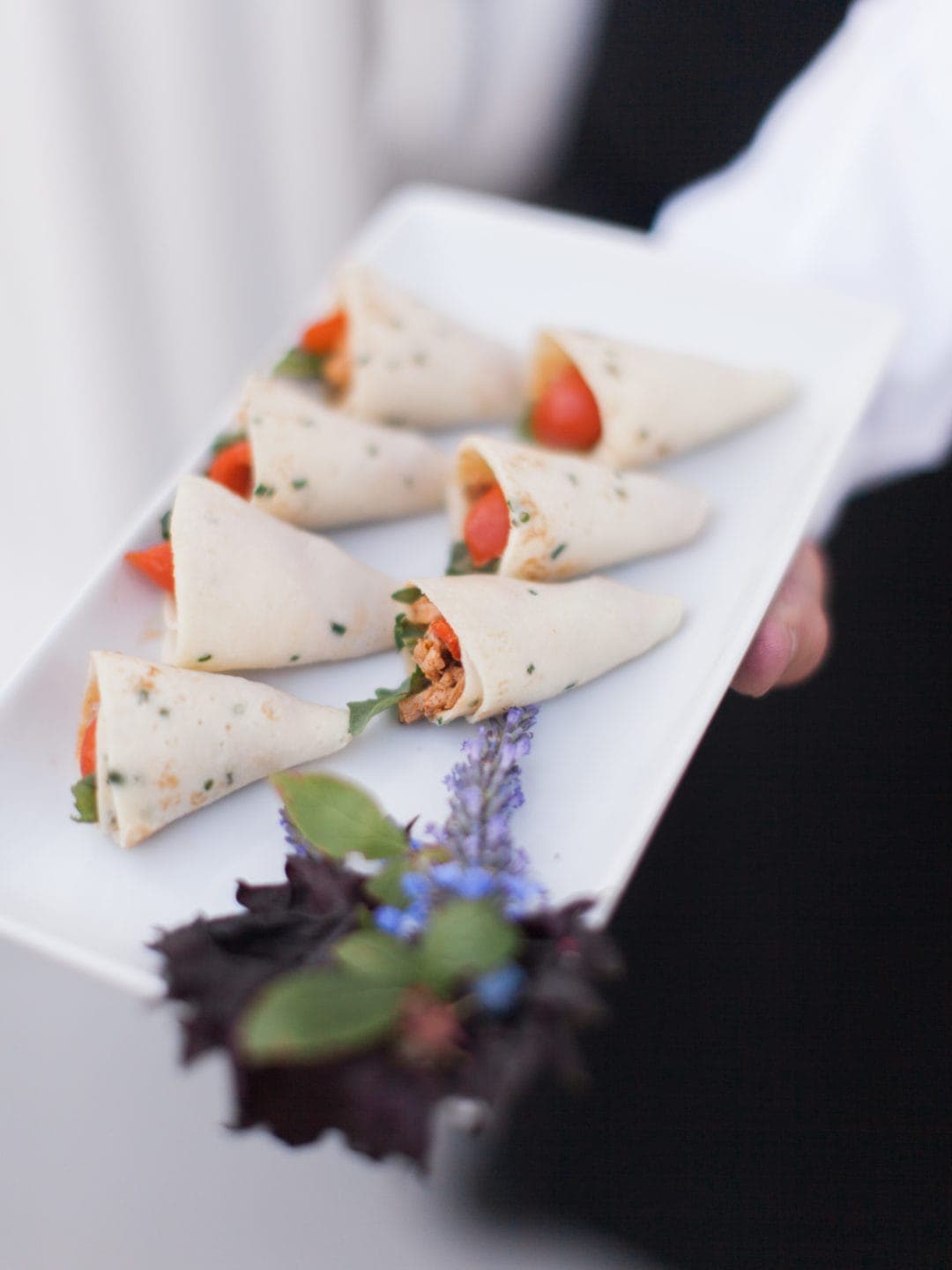 A rectangular white plate with six rolled tortilla wraps filled with vegetables, garnished with greens and purple flowers, held by a person in a white shirt and black vest—perfect for elegant wedding catering in Seattle.