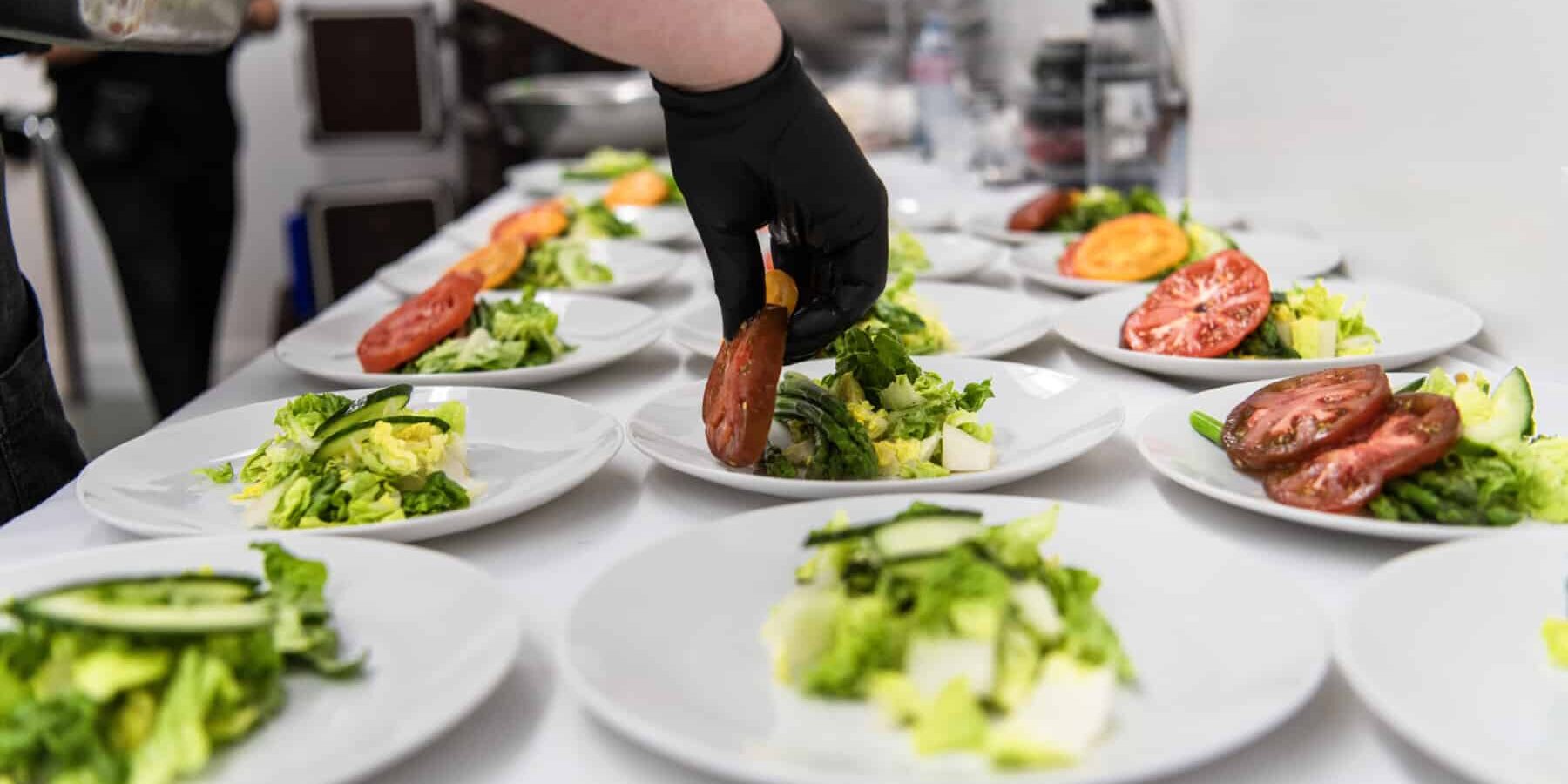 A person wearing a black glove arranges tomato slices on plates of salad lined up on a counter, showcasing the precision and care of wedding catering in Seattle.
