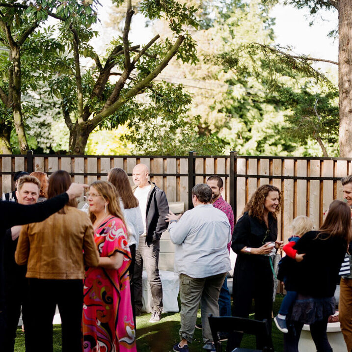 A group of people socializes in a backyard with trees and a wooden fence, some standing and some sitting on outdoor furniture, enjoying delicious wedding and event catering.