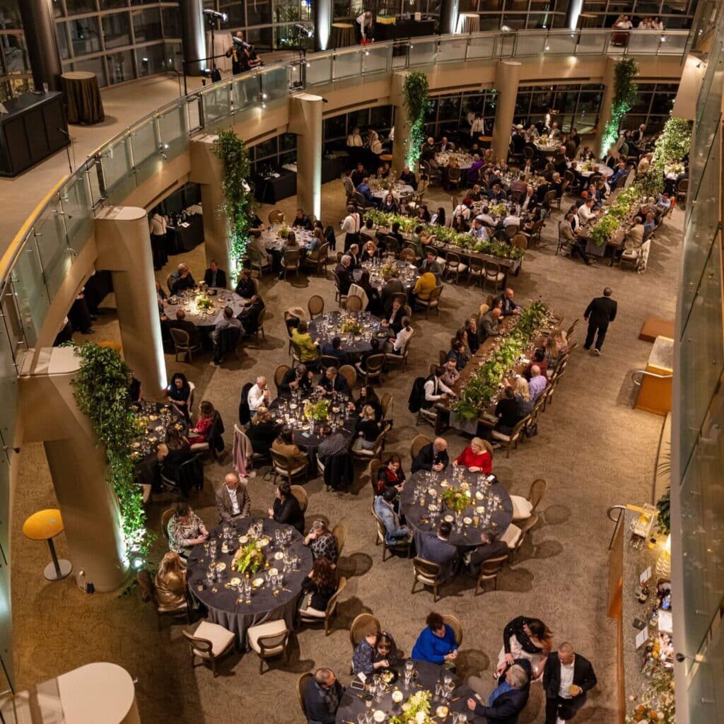 An overhead view of a formal indoor banquet shows guests seated at round tables, dining and conversing in a spacious, well-lit venue with greenery and balcony seating—perfect for catering for fundraisers.