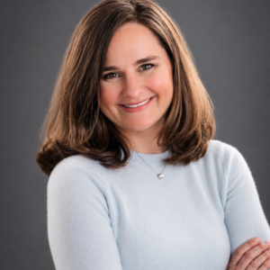 Woman with shoulder-length brown hair, wearing a light blue sweater and a necklace, smiling with arms crossed against a gray background.