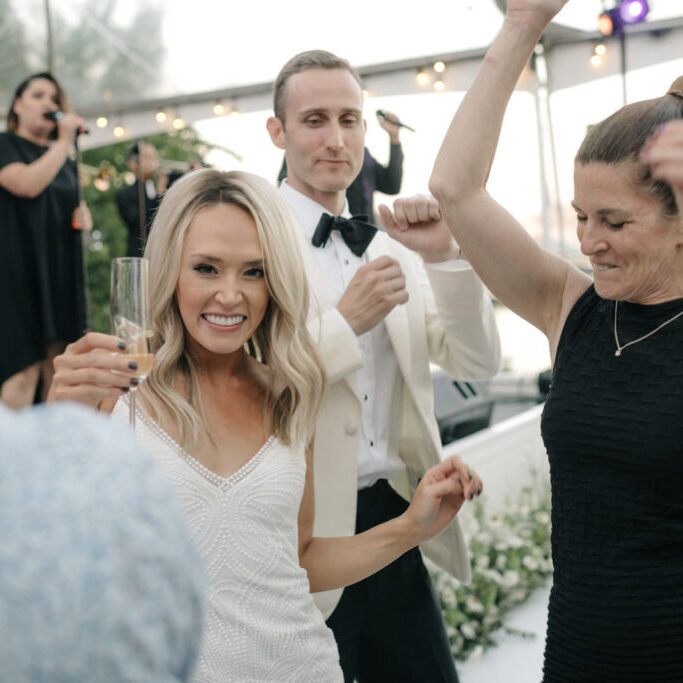 A group of people, including a woman holding a champagne glass, dance and celebrate at a formal event with live music and exquisite wedding and event catering in the background.