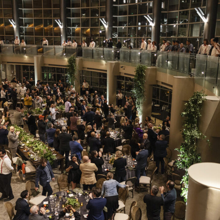 Large group of people attend a formal event in a spacious, modern venue with tables, greenery, and a mezzanine level; many are standing and mingling while enjoying wedding and event catering.