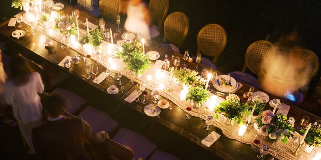 Overhead view of a dimly lit outdoor dinner table with food, drinks, candles, and blurred people—capturing the intimate ambiance of a wedding catering Seattle celebration at night.
