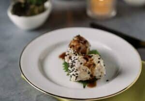 Two pieces of sushi topped with sesame seeds and sauce are served on a white plate, garnished with a green leaf.