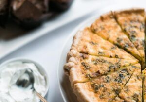 A sliced quiche on a white plate next to a bowl of sour cream with a spoon and a tray of brownies in the background.