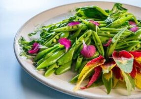 A plate of sliced snap peas garnished with edible flower petals, arranged on a white dish against a light background.