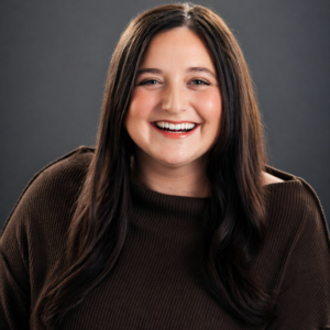 A woman with long dark hair wearing a brown top smiles in front of a plain dark background.