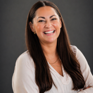 Woman with long dark hair and light blouse smiling, standing against a plain dark background.