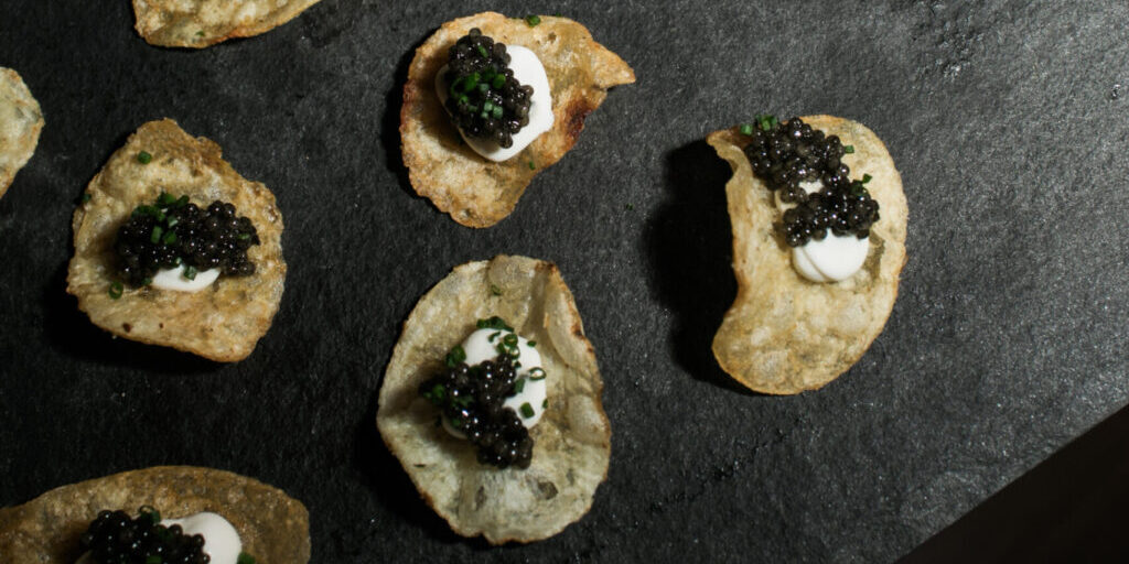 Potato chips topped with sour cream, caviar, and chopped chives arranged on a dark surface.