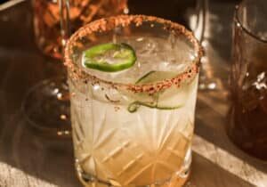 A cocktail in a faceted glass with ice, jalapeño slices, and a chili powder rim, set on a table with other glasses nearby.
