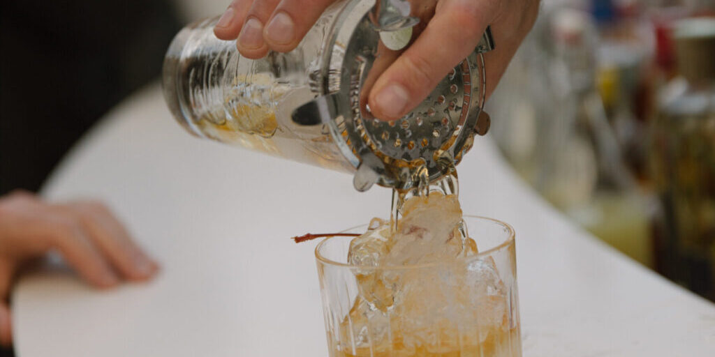 A hand pours a cocktail from a shaker into a glass filled with ice and garnished with a cherry, perfect for party catering.