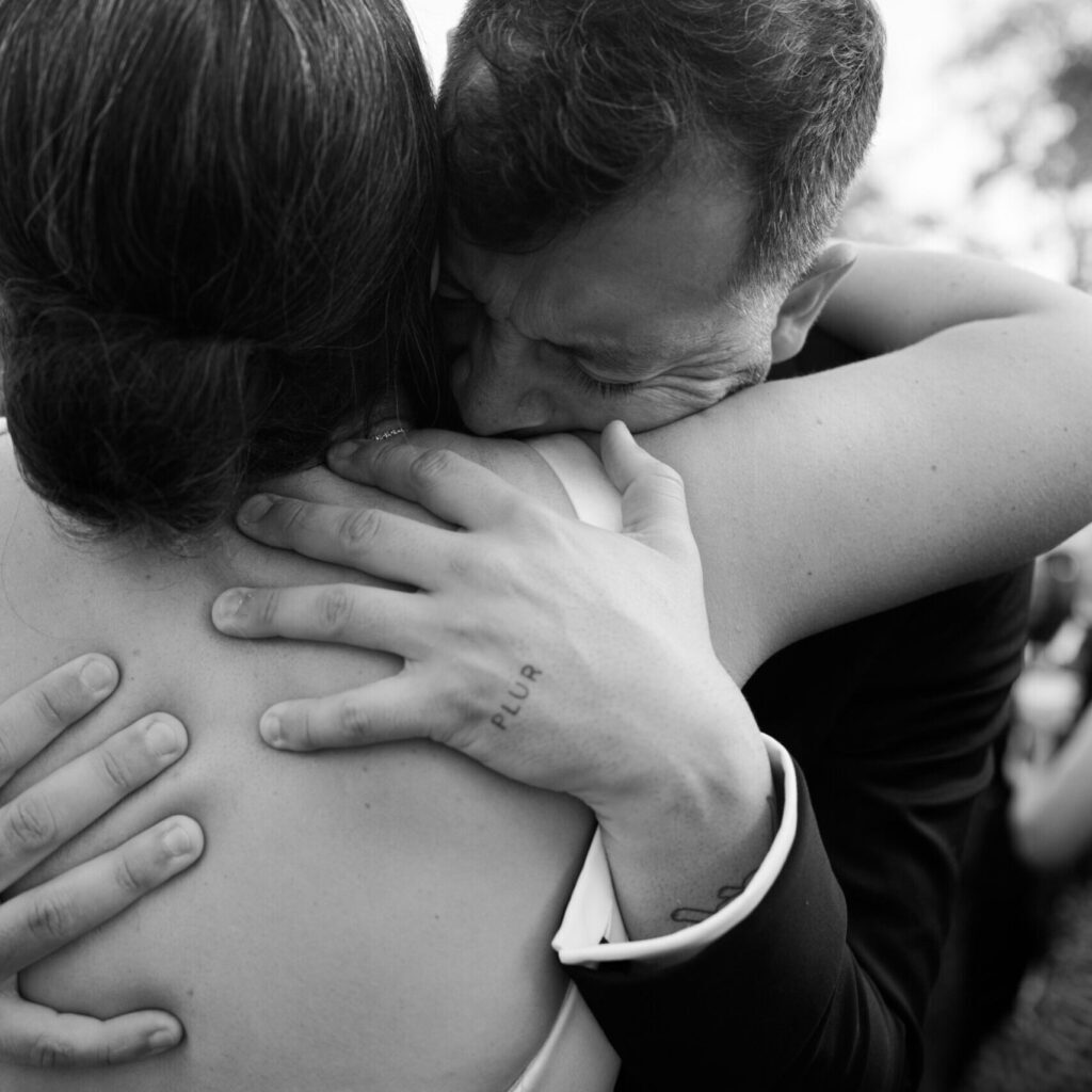 A person embraces another tightly, eyes closed, expressing strong emotion. "PLUR" is tattooed on the hand—a black and white moment as heartfelt as a reunion at top Catering Services in Seattle.