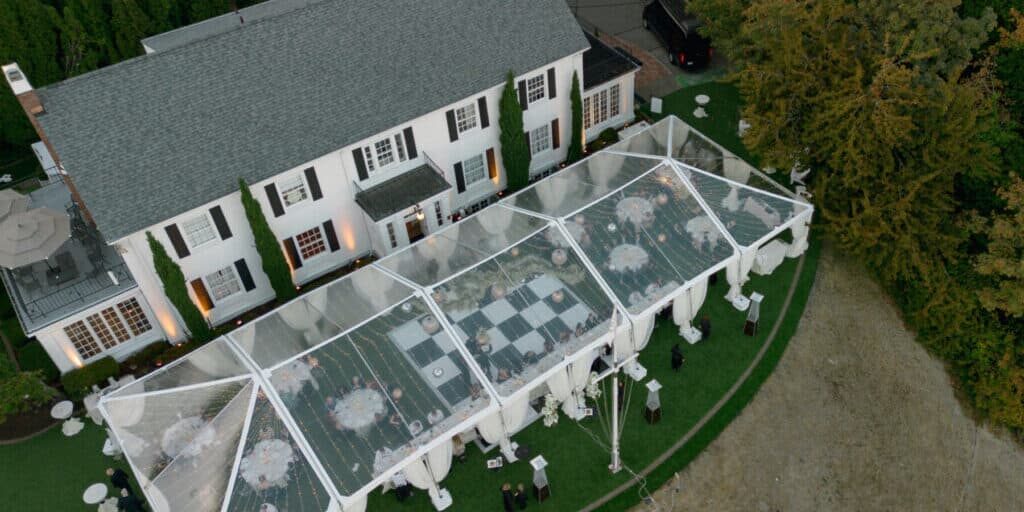 Aerial view of a large white house with a transparent tent set up for an outdoor event, featuring tables, chairs, and a black-and-white checkered dance floor on the lawn.